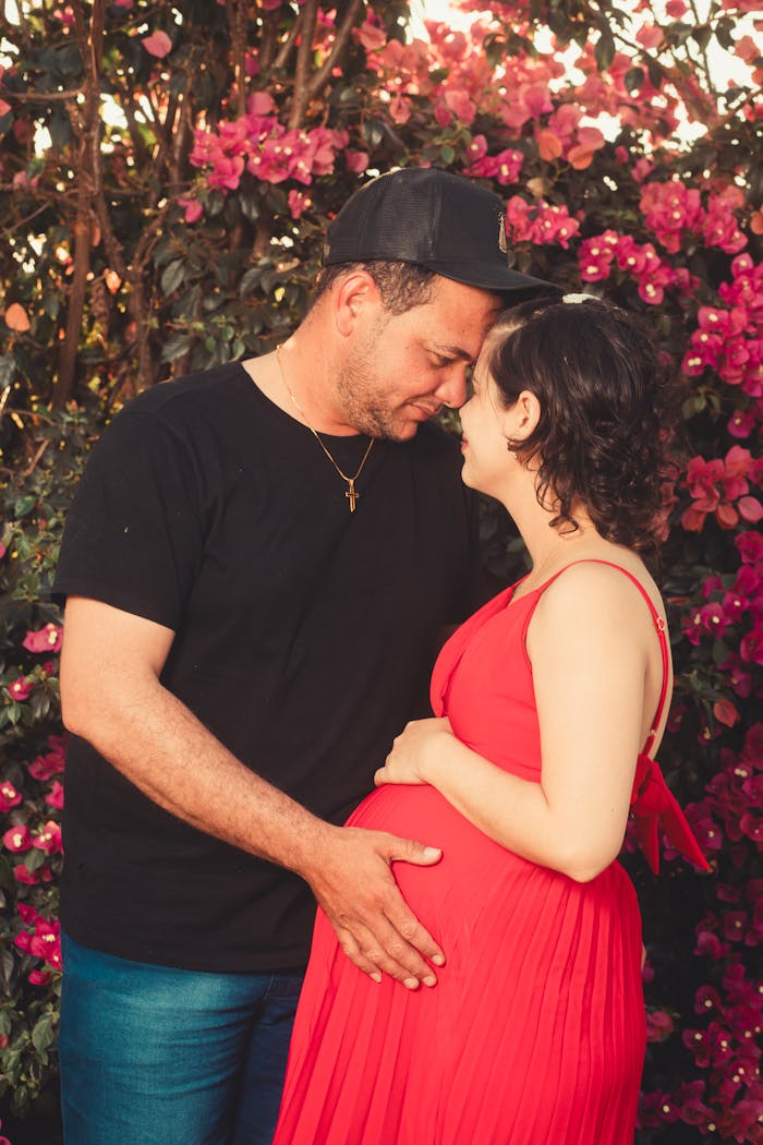 Pregnant couple sharing a tender moment surrounded by vibrant pink flowers outdoors.